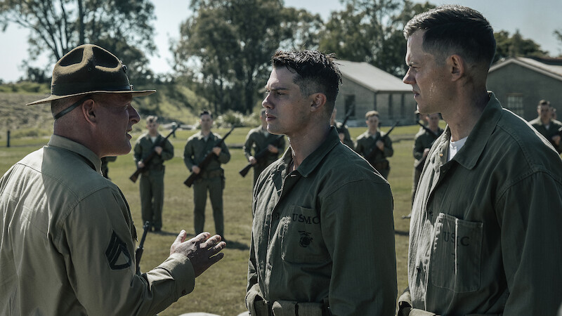 A military officer addresses two soldiers outdoors as other uniformed soldiers with rifles stand in formation behind them on a grassy field with buildings and trees in the background.