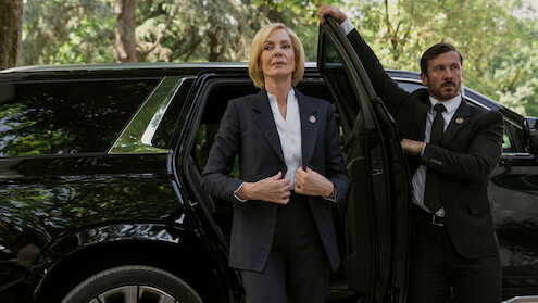 A woman in a suit stands confidently beside a black SUV with a security detail holding the door open, set outdoors with trees and greenery in the background, suggesting a formal or official environment.