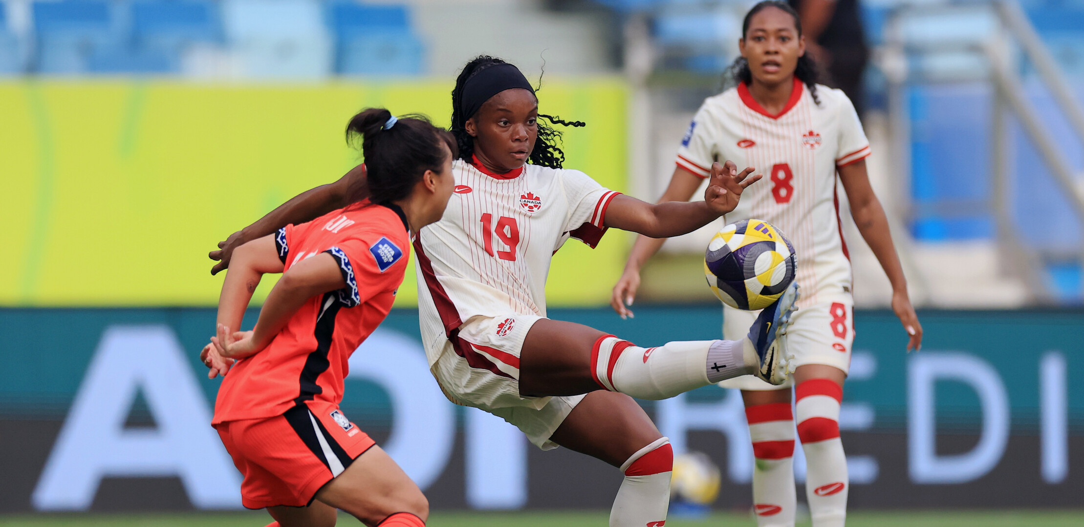 Female soccer players in action during a match, one in a white uniform controls the ball with her foot as opponents and teammates watch, in a stadium with blue seats and a colorful background.