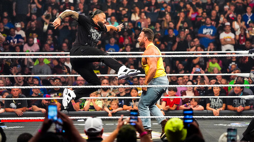 Two professional wrestlers perform a dramatic move in a wrestling ring, surrounded by cheering fans in a packed indoor arena, with several people capturing the action on their phones.