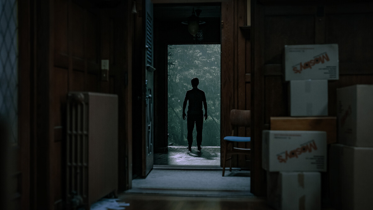 Silhouette of a person standing in a doorway, seen from inside a dimly lit room with moving boxes and wooden paneling, looking out toward a green, misty environment.