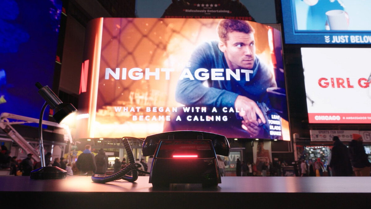 Um grande outdoor digital em uma praça urbana movimentada exibe um anúncio de “O Agente Noturno” com um homem em uma cena de ação. Em primeiro plano, há uma mesa com um telefone retrô e um abajur, cercada por pessoas e pelas luzes da cidade à noite.