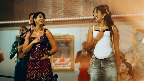 Two women dressed in stylish, colorful outfits smile and dance inside an art gallery, surrounded by other people and framed paintings on the walls, giving a lively and joyful atmosphere.