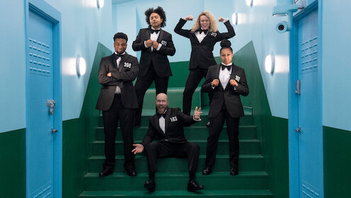 Five people in black suits pose confidently on green stairs in a brightly lit, blue-walled hallway, creating a playful, strong, and upbeat atmosphere.