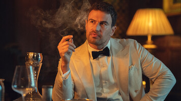 Man in a white tuxedo jacket and black bowtie sits at a dimly lit table holding a cigar, surrounded by wine glasses, a decanter, and a glowing lamp, with smoke swirling in the moody atmosphere.