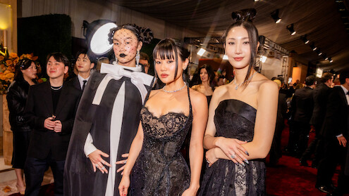 Three people in elegant black and white formal attire pose together at a glamorous indoor event with a red carpet, stylish decor, and other well-dressed guests in the background.