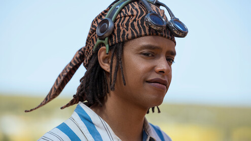 Jacob Romero as Usopp wearing a striped shirt, tiger-patterned headscarf, and goggles stands outdoors in a sunny natural landscape with a blurred background.