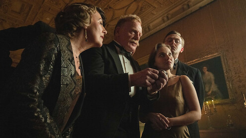 Four well-dressed people in formal evening attire stand close together in an ornate, dimly-lit room with a decorated ceiling and classical artwork, appearing to discuss something intently.
