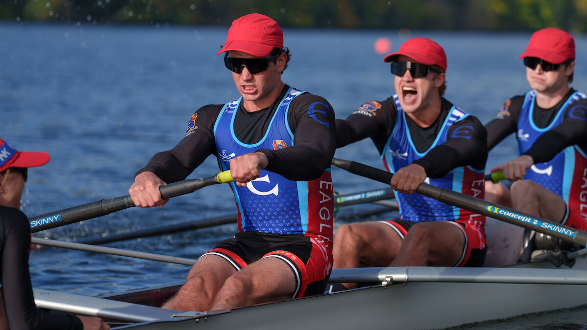 Rowers in matching uniforms and red hats compete in a crew race on a calm river, focused and working together in an outdoor setting with greenery in the background.