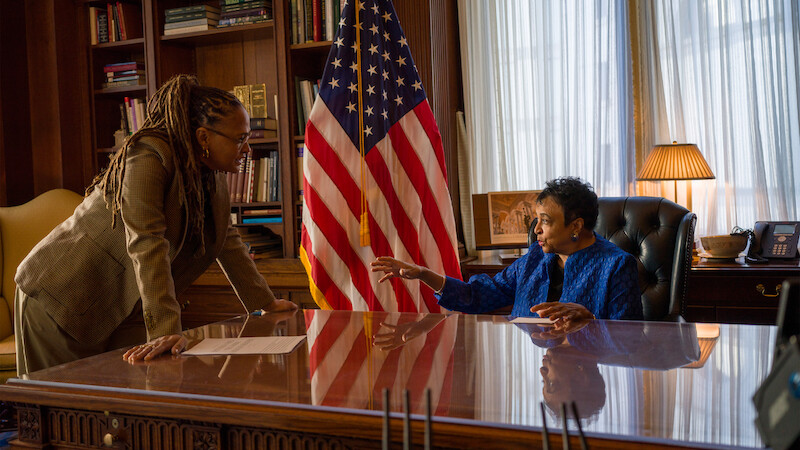 Two women in an office, one standing and one seated behind a polished desk, discuss something. An American flag, bookshelves, and a lamp are in the background, creating a formal, professional setting.