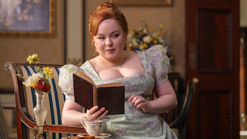 A woman in a floral dress sits in an elegant room with classic decor, reading a book and holding a teacup. Flowers and ornate furniture are in the background, evoking a historical or period setting.