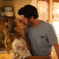 Um casal se beija numa cozinha acolhedora e iluminada pelo sol, com armários de madeira, prateleiras com canecas e frascos, e uma luz natural suave a entrar pela janela.