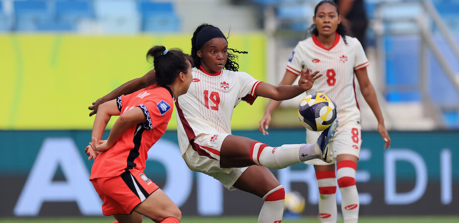 Female soccer players in action during a match, one in a white uniform controls the ball with her foot as opponents and teammates watch, in a stadium with blue seats and a colorful background.