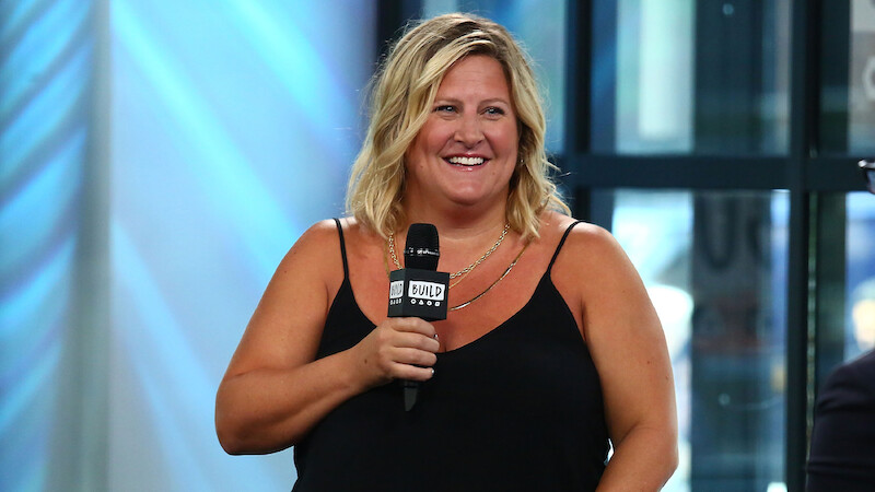 Smiling woman in a black sleeveless top holding a microphone, speaking at a modern indoor event space with large windows, natural light, and a relaxed, positive atmosphere.