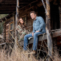 A man and woman sit on the porch of a rustic wooden cabin surrounded by tall grass, wearing casual outdoor clothing. The setting appears peaceful and rural, with natural wooden beams and a weathered structure suggesting a remote location.