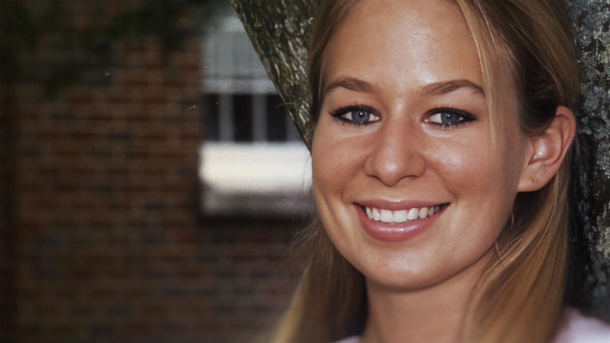 Young woman with long blonde hair smiling, leaning against a tree outdoors, brick building and window blurred in the background, natural daylight.