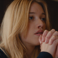 Young woman with long blonde hair indoors, hands clasped in front of her mouth, looking thoughtful, with a small nosebleed visible, soft lighting and neutral background.