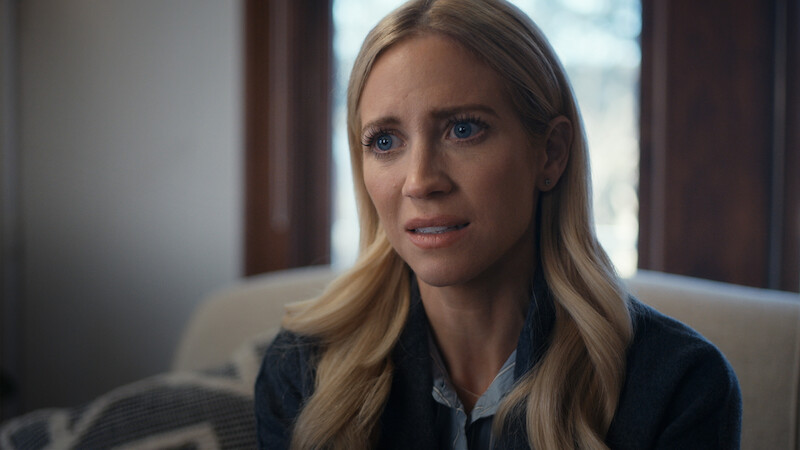 A woman with long blonde hair sits indoors on a couch, looking concerned. She is dressed in a dark sweater over a collared shirt, with soft natural light coming through windows behind her, suggesting a calm, intimate setting.
