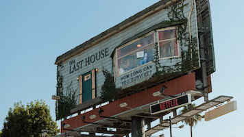 A creative billboard designed as a rundown house with vines, a door, windows, and a sign reading "The Last House" and "How long can you survive?", promoting a Netflix show, set outdoors against a clear blue sky.
