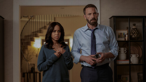 A concerned woman in a blue sweater and a man in a dress shirt and tie stand together in a warmly lit living room, with a staircase and bookshelf in the background, both appearing serious and tense.