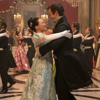 A couple in formal period costumes dances in a grand, elegant ballroom with chandeliers, red drapes, and other couples waltzing in the background during a lavish ball.