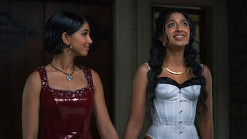 Two young women dressed in formal evening wear stand indoors. One wears a red sequin dress with a heart necklace, the other wears a white corset dress with long hair and shiny makeup. They are holding hands and appear happy.