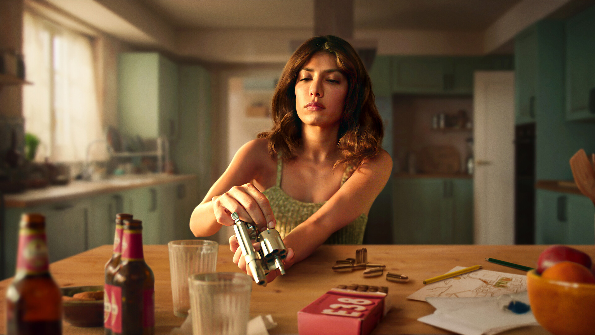 Woman in a kitchen sits at a table with bottles, glasses, and snacks, loading bullets into a revolver, with papers, a red box, and fruit visible, suggesting tension in a domestic setting.