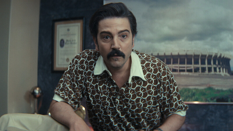 Man with dark hair and mustache in patterned shirt leans forward in an office with framed certificate on wall and large stadium photo in the background.