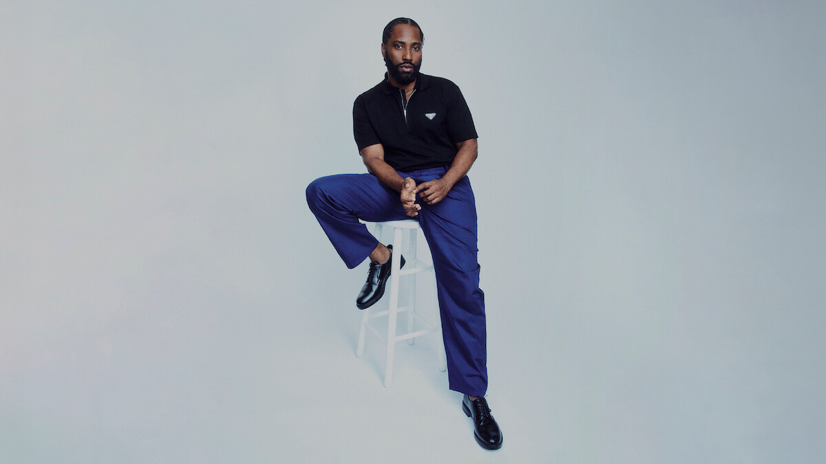 John David Washington sits on a chair and stretches his leg out against a grey background.