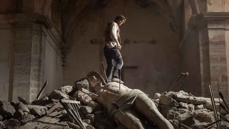 A person stands on rubble inside a damaged, old stone building, with a large broken statue lying among the debris, creating a dramatic and somber atmosphere.