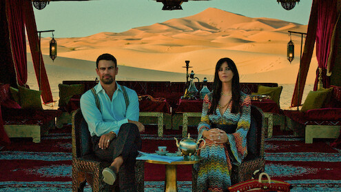 A man and woman sit in an ornate tent with luxurious decor, tea set, and hookah, looking at the camera. The backdrop features vast desert sand dunes under clear sky, lanterns hanging, creating a serene, elegant Middle Eastern ambiance.