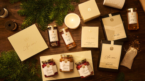 A cozy, warm-toned flat lay of assorted elegantly-packaged gift items, including candles, jars, boxes, and honey, arranged on a wooden table with pine branches, evoking a festive and inviting holiday mood.