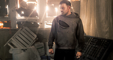 In a still image from the film 'AKA,' actor Alban Lenoir stands in a disheveled room. His hands are at his sides and he wears a ripped gray sweatshirt and black pants. 