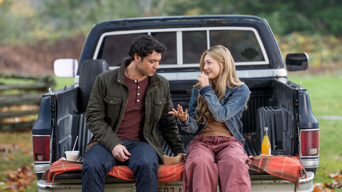 Man and woman sitting on tailgate of a pickup truck in a grassy outdoor setting, talking and eating together, with drinks and food on a blanket, autumn trees in the background.