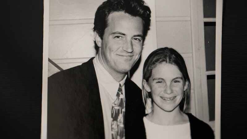 A black and white photo of a man in a suit and a young girl standing side by side, smiling, indoors with a paneled wall and window in the background.