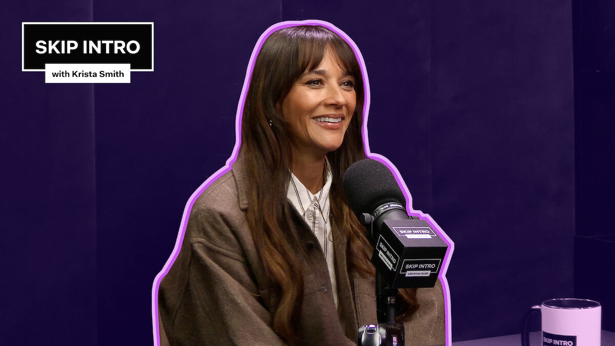 Rashida Jones, sorrindo, sentada diante de um microfone, com um fundo roxo-escuro.