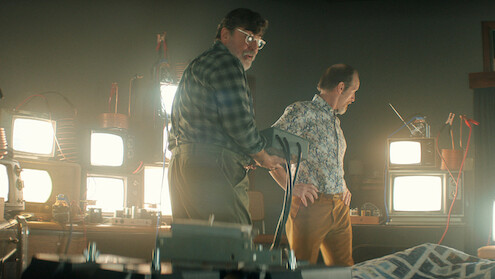 Two men in a dimly lit room filled with old televisions and electronic equipment, one holding a device, standing beside a bed with a patterned blanket, suggesting an experimental or scientific setting.