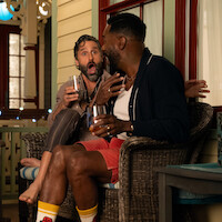 Two men sitting on a porch at night, laughing and talking while holding drinks, with one looking surprised; cozy, relaxed atmosphere with decorative lights and porch furniture.