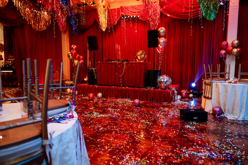 Um salão em tons de vermelho onde uma festa acabou de terminar tem papel picado e adereços brilhantes por toda parte