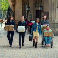 Five people carrying boxes and bags, including one in a wheelchair, walk together outside a historic stone building with arches and lanterns, suggesting a college or university move-in day.