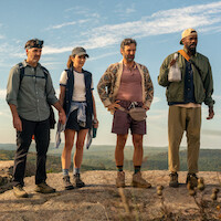 Four hikers in outdoor gear stand on a rocky hilltop with forested landscape and blue sky in the background, prepared for a nature trek or adventure.
