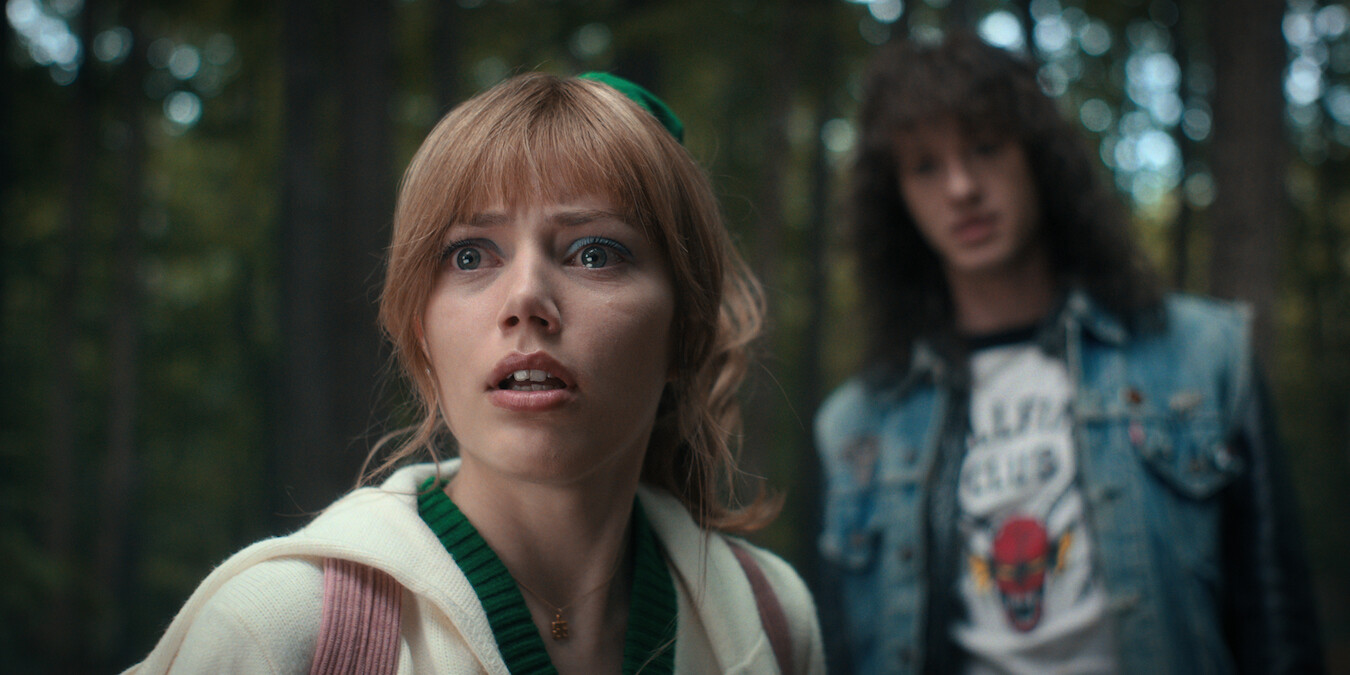 Chrissy Cunningham (Grace Van Dien), looks startled in the foreground while Eddie Munson (Joseph Quinn), stands blurred in the background in a wooded area.