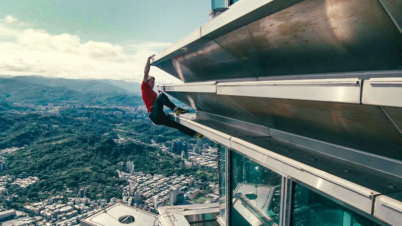 Alex Honnold in a red shirt climbing the exterior of a tall skyscraper above a city, with mountains and buildings visible in the distant background under a partly cloudy sky.