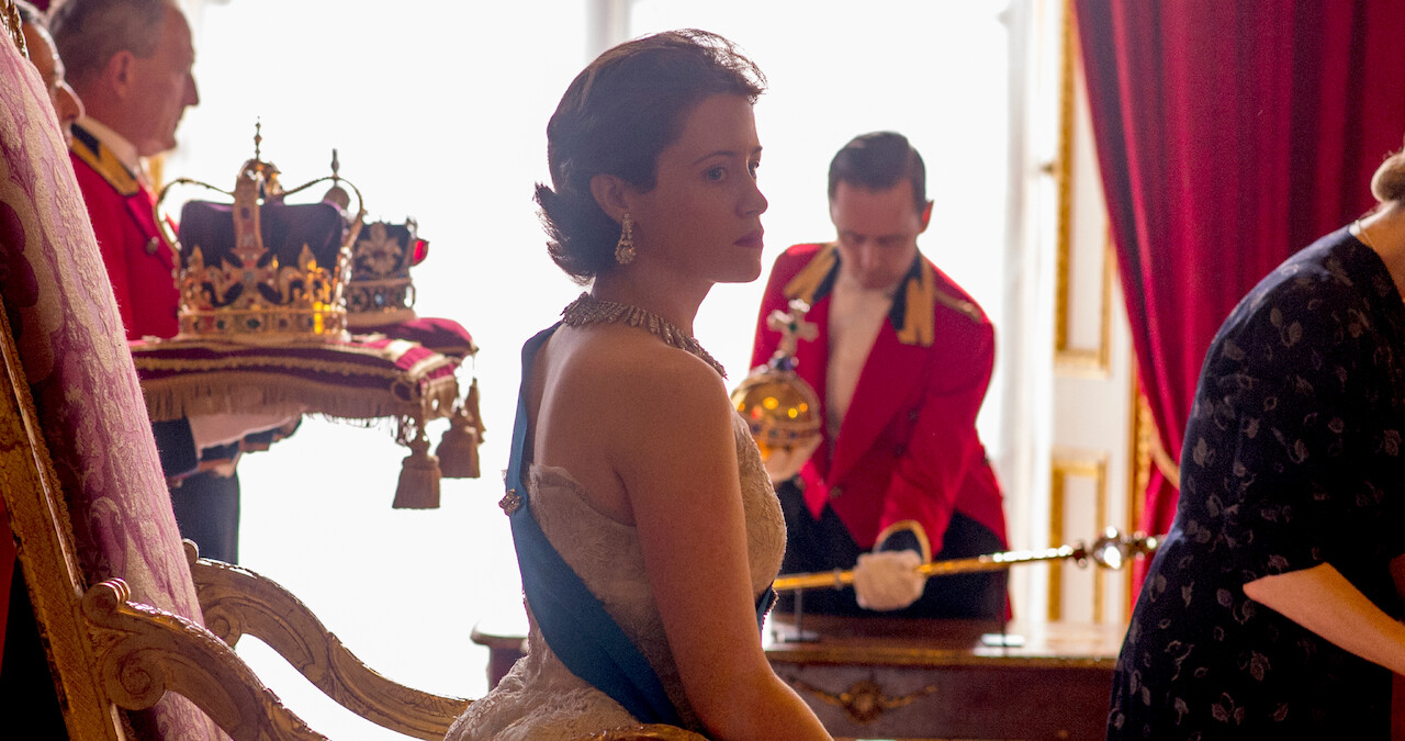 Claire Foy as the Queen in The Crown. She's sitting behind a desk with the royal crown behind her
