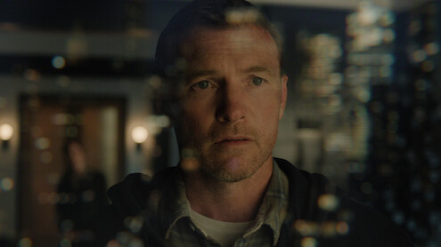 A man stands indoors, looking intently through a glass wall with light reflections; the environment is dimly lit with modern decor, and a blurred figure is visible in the background.