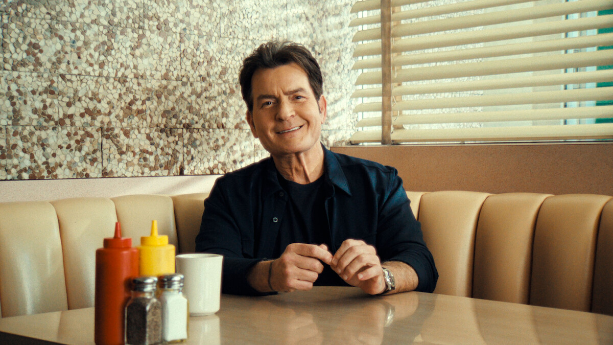 A smiling Charlie Sheen sits in a diner booth.