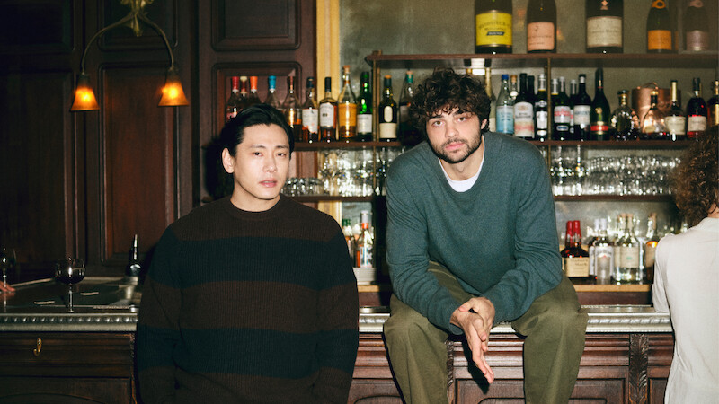 Teo Yoo leans against, and Noah Centineo sit atop the bar looking directly to camera.