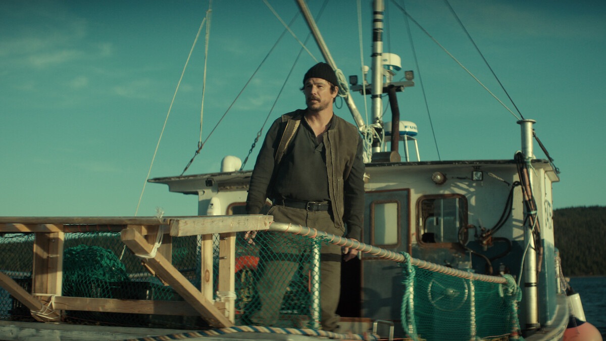 A man wearing a beanie stands on the deck of a fishing boat surrounded by nets and wooden railings, with the sea and distant land visible under a clear sky.