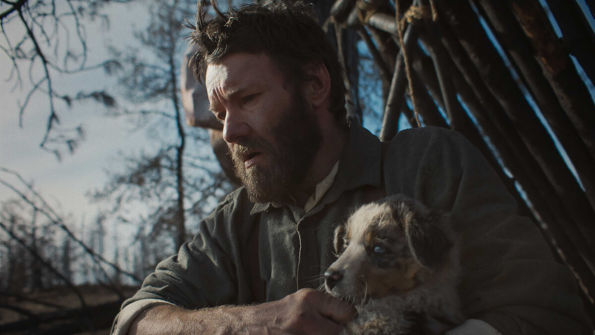 A bearded man sits outdoors in a rustic, wooden shelter, holding a small dog in his arms. The environment looks cold and barren, evoking a somber, contemplative mood as sunlight filters through the sparse trees.