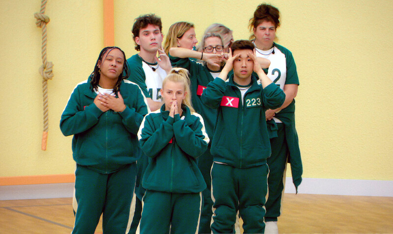 A group of people in green tracksuits stands indoors against a yellow wall, expressing anxiety and tension, suggesting participation in a competitive or high-stakes event.